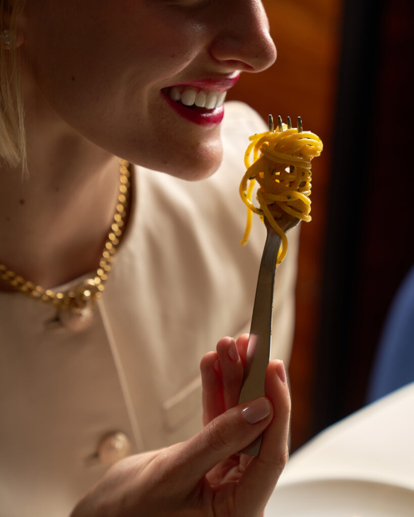 a woman with red lips and gold jewelry about to take a bit of spaghetti