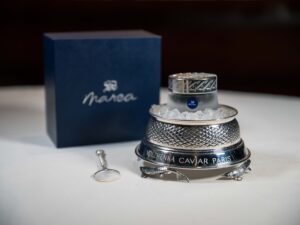 marea volzhenka caviar - a glass jar of marea branded caviar sits on a bed of ice in a silver serving tray. propped up on the left is the lid of a box: navy blue with a silver marea logo. in front of the box lid, a silver and mother of pearl serving spoon lays on the white tablecloth