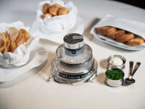 marea volzhenka caviar - a glass jar of marea branded caviar sits on a bed of ice in a silver serving tray. it is displayed surrounded by various accoutreuments - gougeres, chips, brioche, chives, and crème fraîche