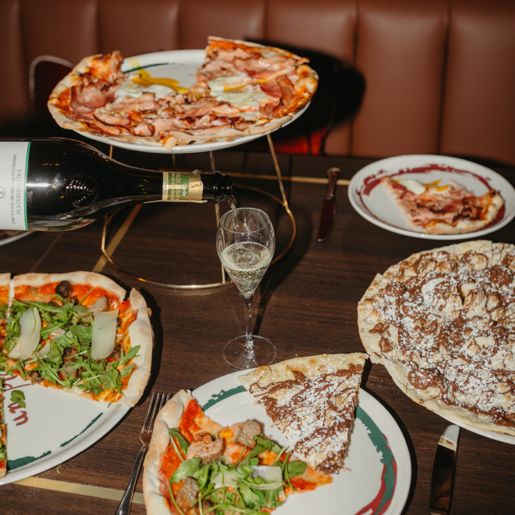 wine being poured into a glass on a table full of various pizzas at crazy pizza