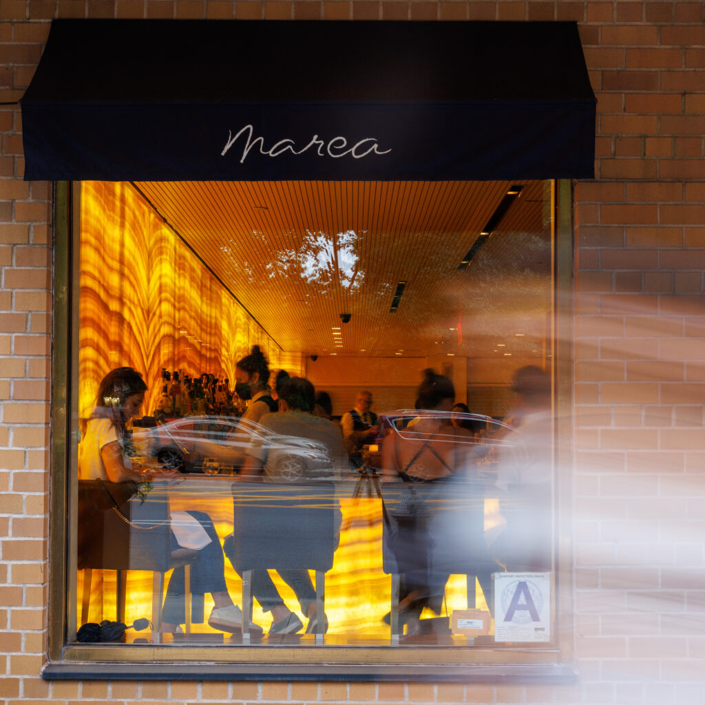 the exterior of marea: a dark blue awning bearing the marea logo is seen over a window through which we can see into a busy bar, lit by the famous honey onyx