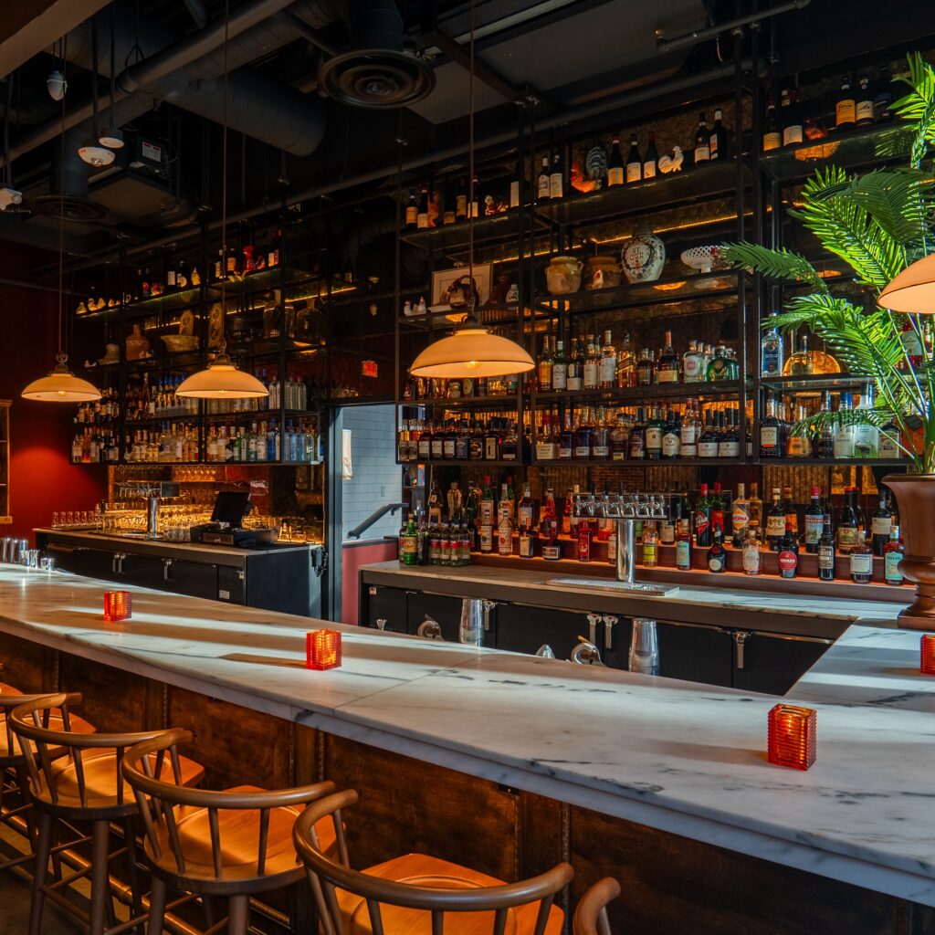 the bar at cucina morini - lit with a warm light, shelves go to the ceiling full of bottles behind a white marble bar, with wooden stools
